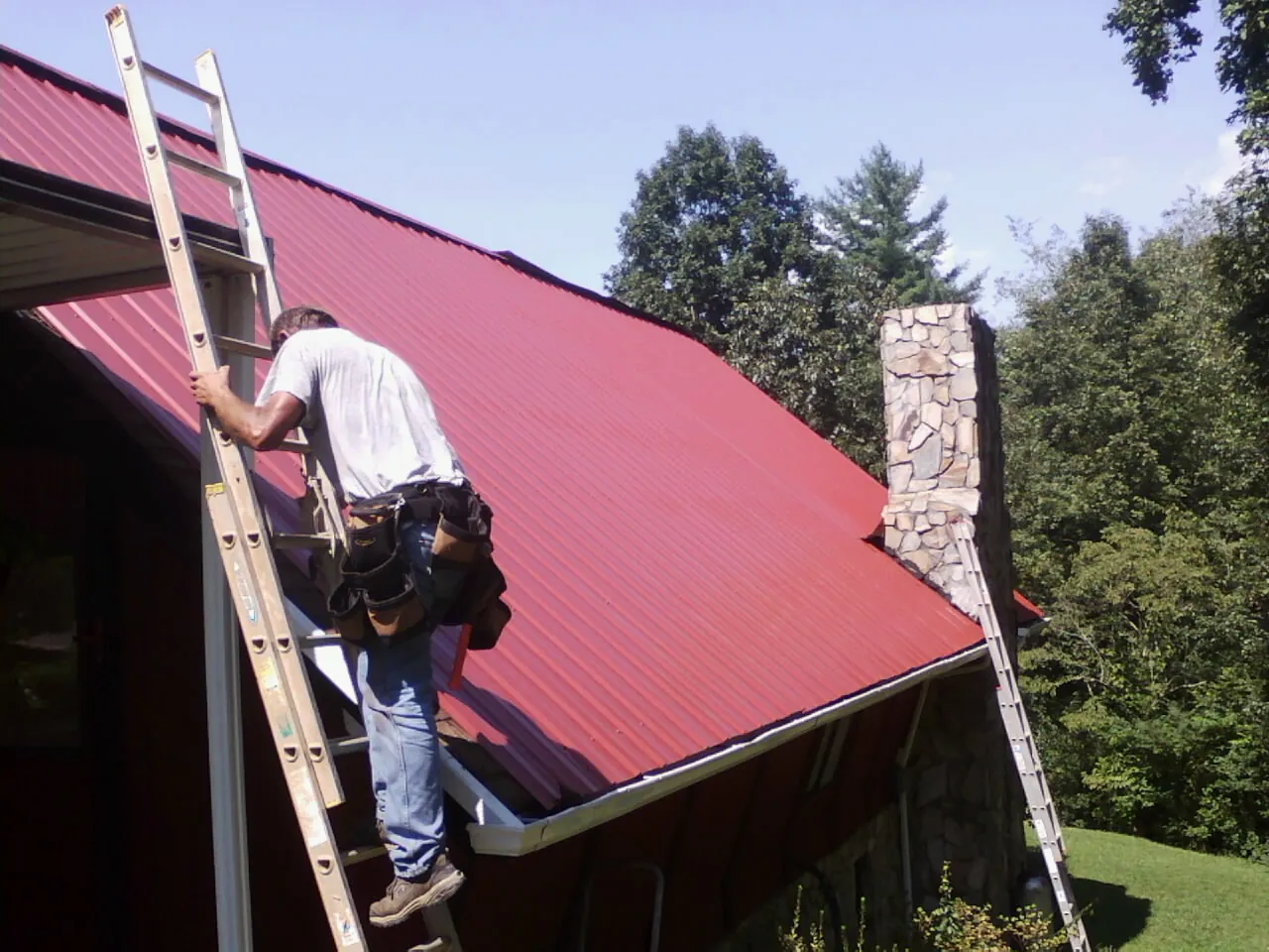 Roofer on a ladder beside a red metal roof and stone chimney — Soffit & Fascia Repair by Roofing Brothers in Selma